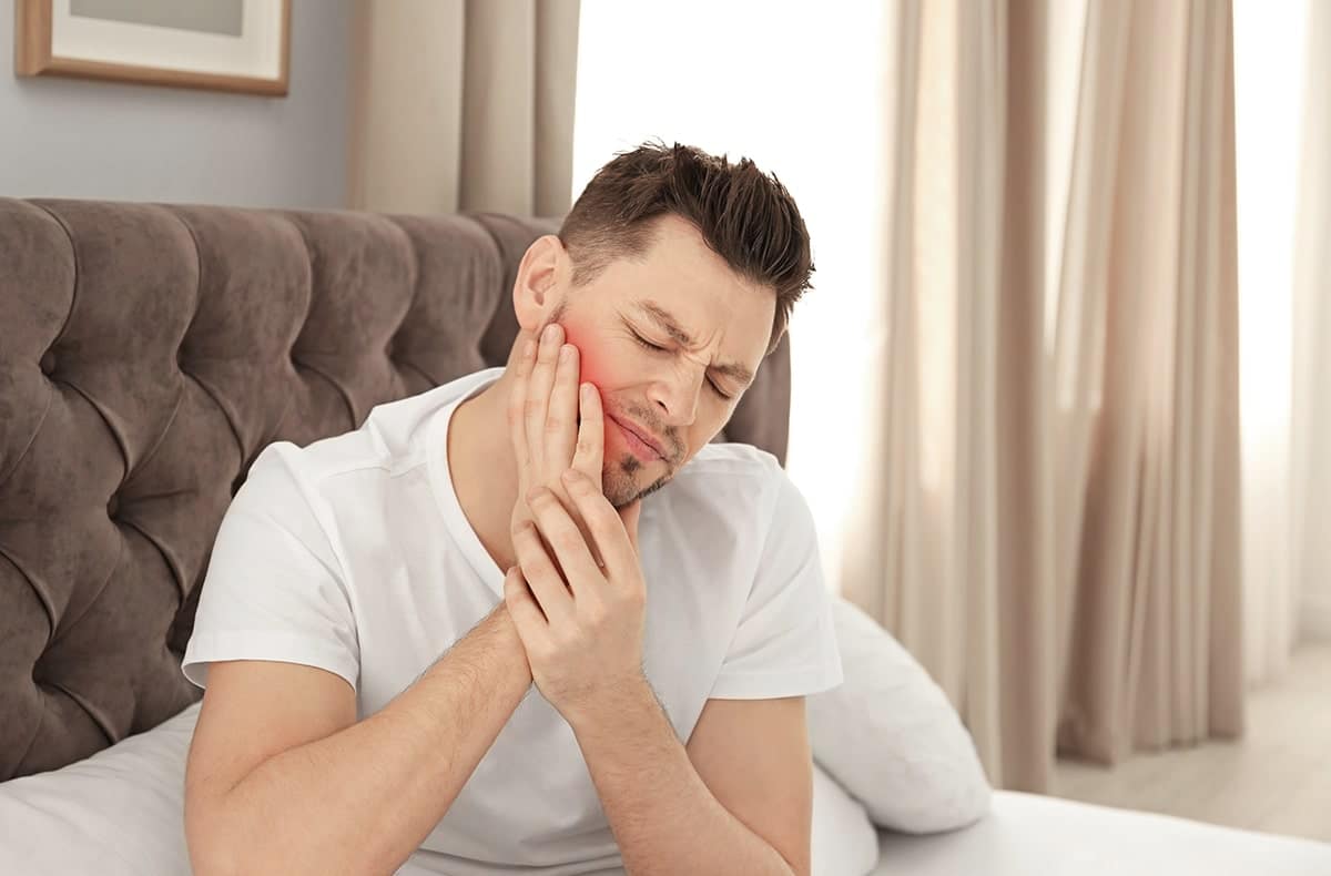 man just waking up from bed holds the side of his face due to jaw pain